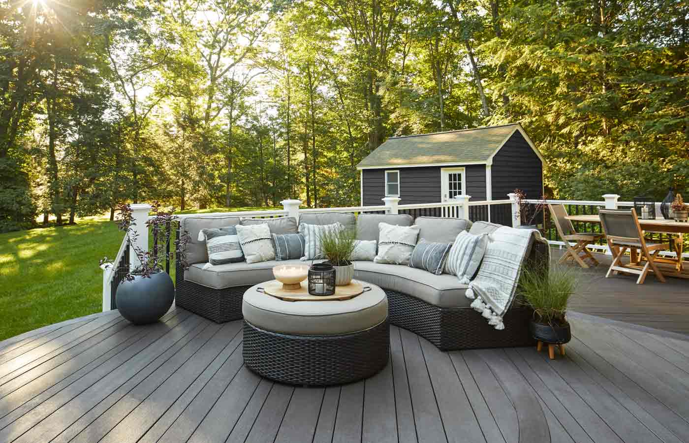 TimberTech-Dark-Hickory-Boston-Vintage-Collection-AZEK-Decking-multi-width-inspiration-gallery-1 Modern outdoor deck with a curved sectional sofa, round ottoman, potted plants, and a dining table set, surrounded by trees and a small shed in the background.
